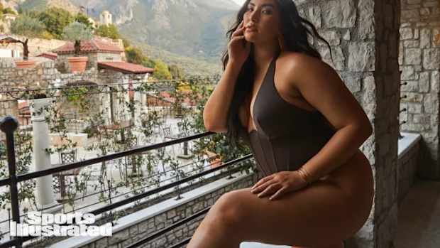Yumi Nu leans on a railing overlooking the city, wearing a olive one-piece swimsuit.